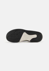 Semelle de chaussure en caoutchouc noir avec adhérence texturée, semelle intermédiaire blanche et rainures distinctes pour la traction. Logo embossé sur le côté.