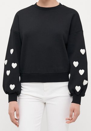 Pull noir à manches longues avec des motifs de cœurs blancs sur les manches, porté avec un pantalon blanc, vue de face.
