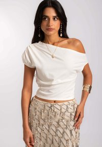 Haut blanc, décolleté bardot avec des manches courtes bouffantes, associé à une jupe en crème perlée ornée d'accents argentés et dorés ainsi que d'un motif géométrique.
