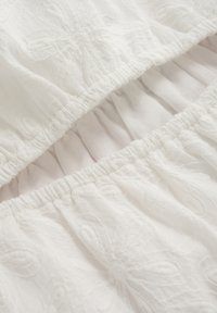 Abito bianco con motivi floreali ricamati e vita arricciata, con tessuti sovrapposti che creano un aspetto textured.