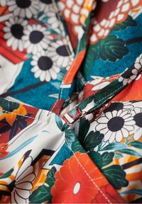Tissu à motifs floraux avec des couleurs vives rouge, orange et bleu-vert. Texture lisse avec une couture visible et un détail en métal.