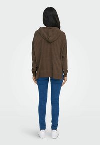 Brun ribbad hoodie med en söm på baksidan och en huva, kombinerad med blå tajta jeans och vita sneakers. Enfärgad bakgrund.