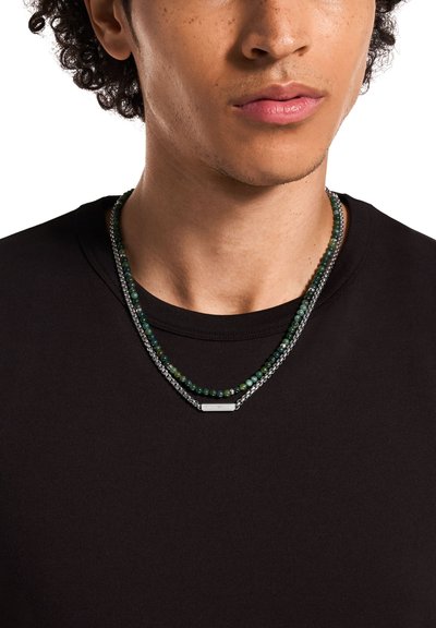 Collier superposé avec perles vertes et chaîne en argent, agrémenté d'un accent métallique rectangulaire ; porté sur une chemise noire.