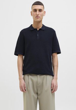 Polo shirt texturé bleu marine avec une patte de boutonnage à trois boutons, des manches courtes et une coupe décontractée, associé à un pantalon beige clair à plis.
