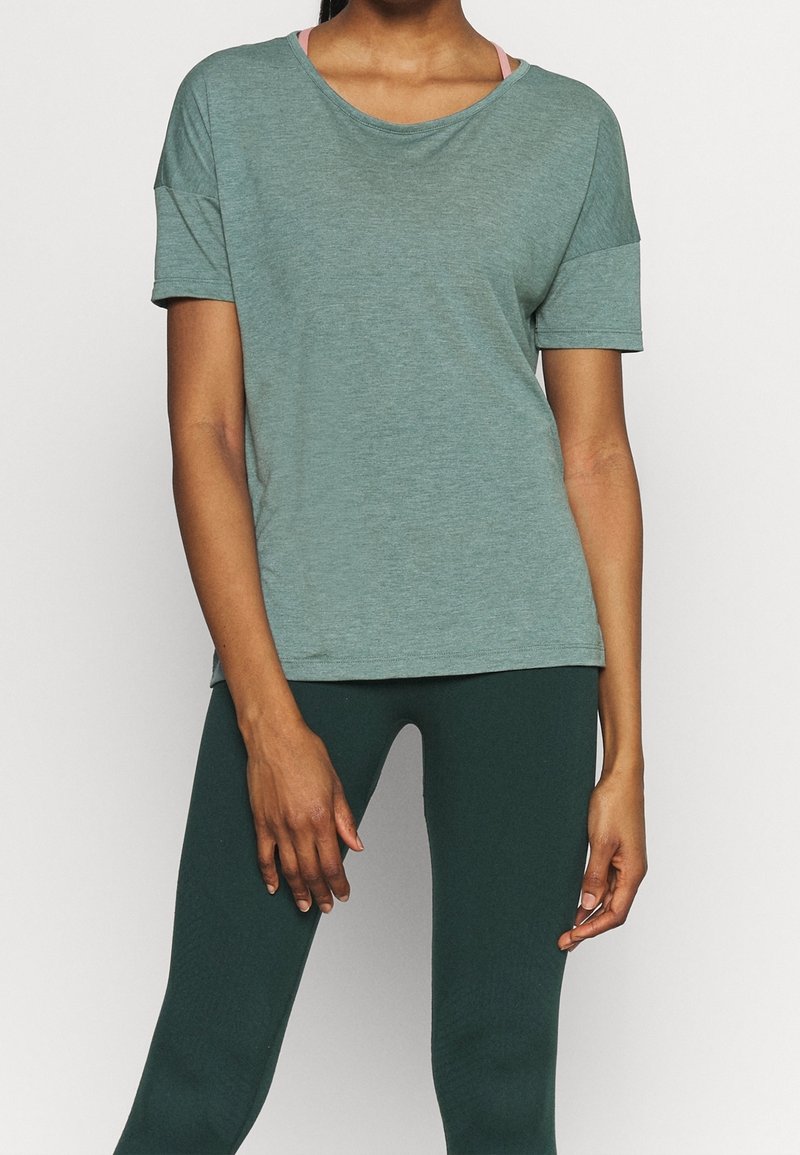 T-shirt à manches courtes, couleur vert doux, avec un col arrondi, une coupe décontractée et des manches retroussées. Porté par-dessus des leggings vert foncé.