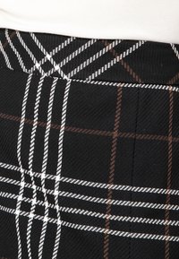 Musta ruudullinen hame, jossa on teksturoitu kangas ja valkoisia sekä ruskeita viivoja, jotka luovat risteäviä kuvioita. Yksityiskohtainen vyötärö, jossa ei ole näkyvää mekanismia.