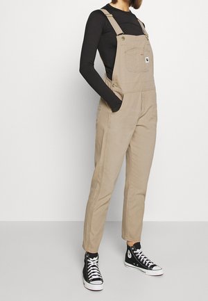 Person iført beige overall med en sort langærmet trøje og sorte høje sneakers, stående med hænderne i lommerne.
