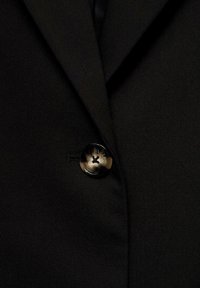 Sort blazer med en stor, rund, tofarvet knap med et korsdesign. Stoffet ser glat ud med en skræddersyet pasform og skarpe revers.