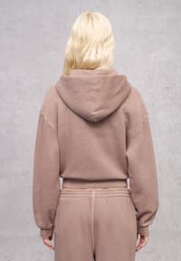 Sudadera marrón con una gran capucha, puños y dobladillo acanalados. Presenta texto en relieve en la parte posterior. Tejido suave y texturizado, combinado con pantalones deportivos a juego.