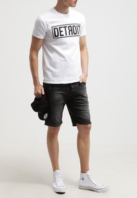 Hombre con camiseta blanca de "DETROIT", pantalones cortos de mezclilla negros, zapatillas altas blancas, sosteniendo una gorra negra en la mano izquierda, de pie contra un fondo liso.