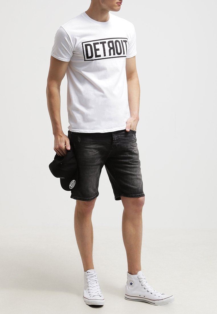 Hombre con camiseta blanca de "DETROIT", pantalones cortos de mezclilla negros, zapatillas altas blancas, sosteniendo una gorra negra en la mano izquierda, de pie contra un fondo liso.