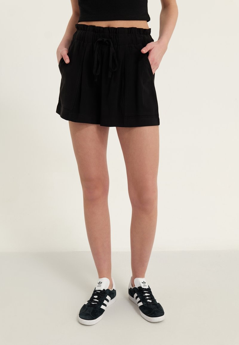 Shorts en tissu noir avec une taille élastique et un cordon de serrage. Dotés de poches latérales et d'une coupe ample. Associés à des baskets noires et blanches.