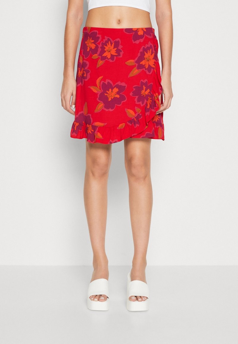 Femme portant une jupe portefeuille rouge avec de grands motifs floraux violets et orange et des sandales compensées blanches, visible du ventre jusqu'aux pieds.