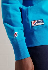 Sudadera turquesa con puños acanalados, que presenta un pequeño logotipo rojo y azul en la manga y una etiqueta de parche en el cuerpo. Textura suave.