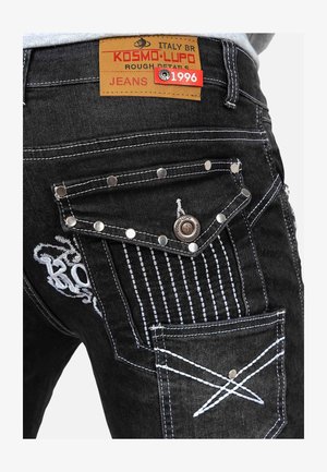 I jeans in denim nero presentano tasche posteriori stilizzate con hardware argentato, cuciture bianche e motivi ricamati. Etichetta in pelle visibile con dettagli del marchio.