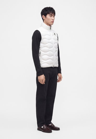 Hombre de pie, lleva un chaleco blanco acolchado sin mangas, una camisa negra de manga larga, pantalones negros y mocasines marrón oscuro sobre un fondo blanco.