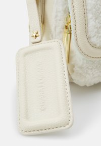 Jasnobeżowa skórzana torba o teksturowej powierzchni, złotym okuć i odpinanym prostokątnym znaku. Posiada puszysty biały akcent.