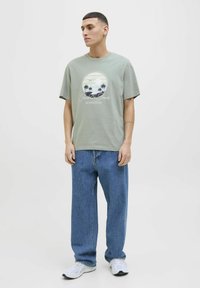 Camiseta de algodón color verde oliva con un gráfico circular que presenta un atardecer, palmeras y una marca; combinada con unos vaqueros azules holgados y zapatillas blancas.