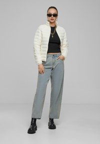 Veste en crème légèrement rembourrée, crop top noir, jean droit bleu clair, et bottes à talons épais noires. Les accessoires incluent des lunettes de soleil et des colliers superposés.