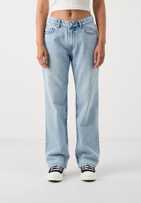 Ανοιχτό μπλε τζιν παντελόνι φαρδύς γραμμής με ψηλή μέση, διαθέτοντας κλασικές τσέπες και ραφές, συνδυασμένο με μαύρα και λευκά αθλητικά παπούτσια.