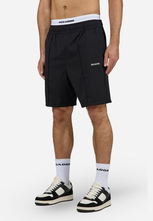 Man draagt zwarte Pegador-shorts, witte Pegador-sokken en zwart-witte sneakers, staand tegen een effen achtergrond.