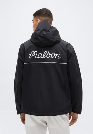 Homme portant une veste noire à capuche avec l'inscription blanche "Malbon" et une ligne horizontale au dos, associé à un pantalon blanc, debout devant un fond uni.