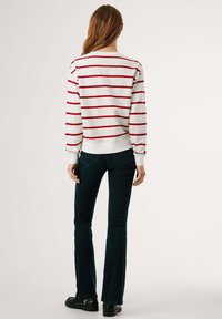 Suéter blanco con rayas horizontales rojas, cuello redondo, puños y dobladillo acanalados. Combinado con vaqueros oscuros acampanados y zapatos negros.