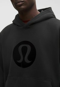 Fekete kapucnis pulóver, nagy, kör alakú, domború logóval. Az anyag puha megjelenésű, texturált felülettel. Kanguru zseb található rajta.