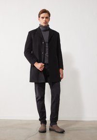 Abrigo de lana negro con un solo botón, suéter de cuello alto gris, pantalones de mezclilla oscuros y botas grises, de pie frente a un fondo blanco sencillo.