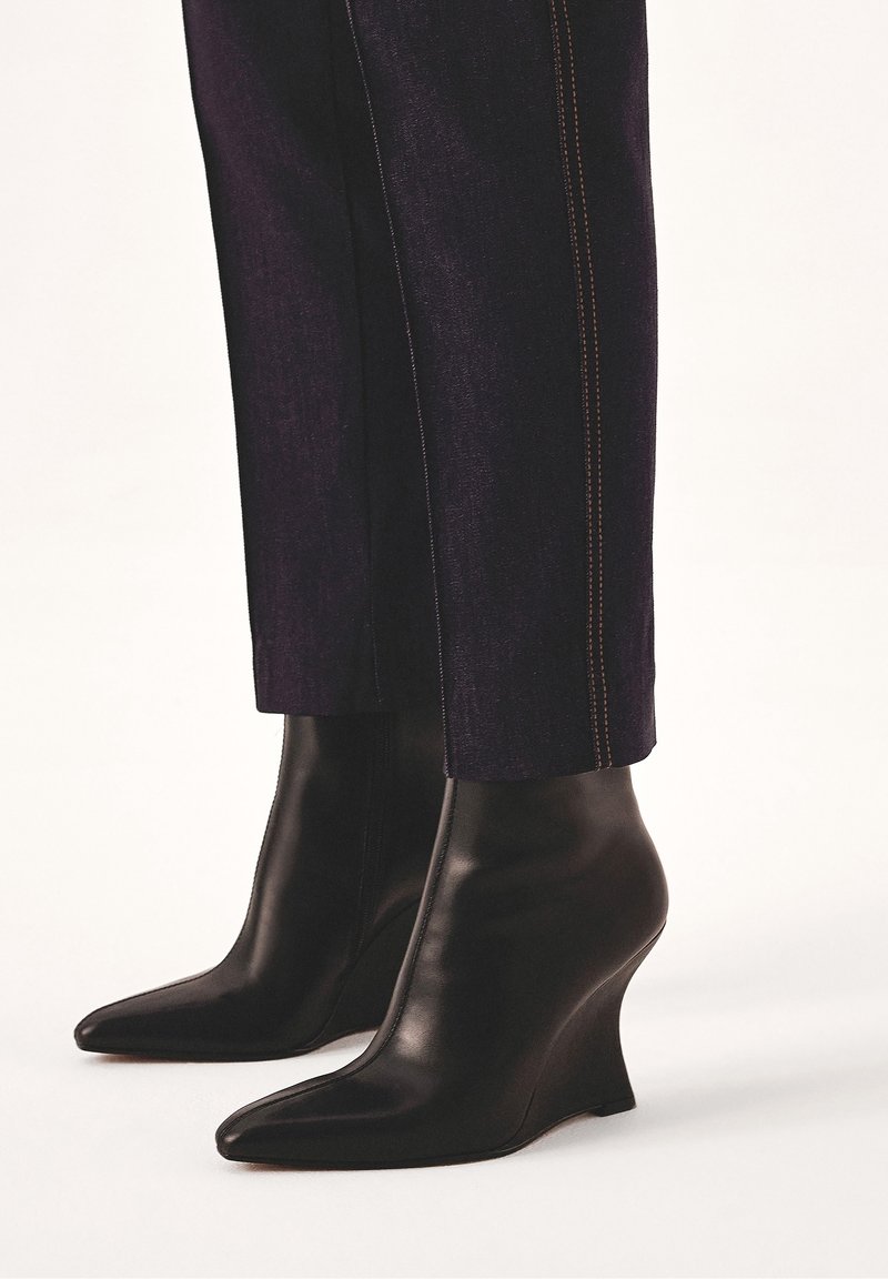 Bottines à talon compensé en cuir noir portées avec un pantalon foncé avec couture latérale visible, vues de mi-mollet vers le bas.