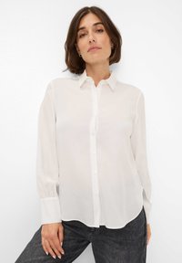 Camicia bianca, trasparente, con colletto, maniche lunghe e orlo curvo. Il tessuto appare leggero e liscio, esaltando il suo design minimale.