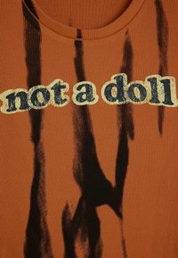 Roostepunane soonikkoes T-särk, millel on tekst "not a doll" kreemikas ja laivahallis värvitoonis ning musta abstraktse värvilainetusega kangal.