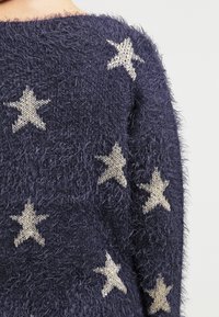 Maglione blu navy a maglia con una texture morbida, caratterizzato da motivi a stella color crema. Design a boxy con un collo ampio.