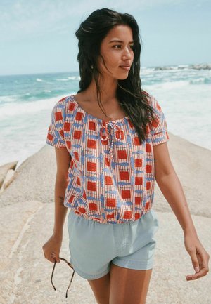 Hosszú, sötét hajú nő, mintás blúzt és világoskék rövidnadrágot viselve, köves úton sétál az óceán partján, miközben napszemüveget tart.