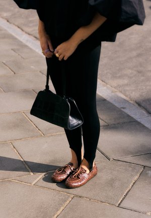 Persona che indossa leggings neri, tiene una borsa nera e indossa scarpe in pelle marrone stile mocassino su un marciapiede lastricato in pietra.
