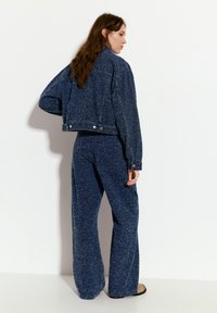 Modrá strukturovaná džínová bunda s širokými rukávy a zkráceným střihem, doplněná volnými kalhotami ve stejném stylu. Obsahuje detaily se zacvakávacími knoflíky.