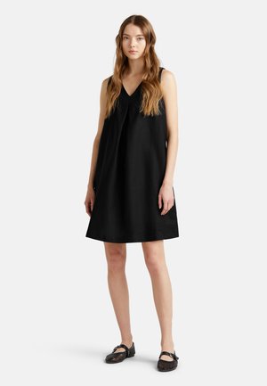 Robe noire sans manches avec un col en V et une silhouette évasée, faite d'un matériau ressemblant à du lin. Associée à des chaussures plates tissées noires.