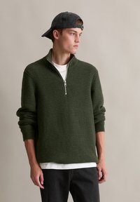 Grüner Strickpullover mit halber Reißverschluss-Kragen, strukturiertem Muster und gerippten Bündchen, kombiniert mit einem weißen T-Shirt und schwarzen Jeans.