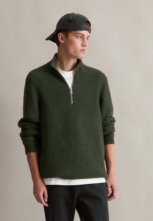 Knit green pullover with half-zip collar, textured pattern, and ribbed cuffs, paired with a white T-shirt and black jeans.