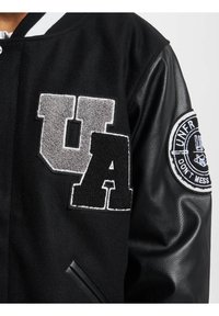 Veste varsity noire avec des patchs en feutre texturé gris et noir "UA", des manches en cuir et un logo brodé rond sur le bras droit.
