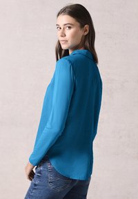 Langärmliges Hemd aus leuchtend blauem Stoff, mit Kragen und abgerundetem Saum. Trägt es mit blauen Jeans, die eine glatte Textur und Passform zeigen.