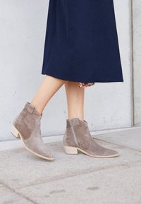 Stivaletti in camoscio grigio alla caviglia con punta affilata, tacco basso impilato e cerniera laterale, indossati con una gonna blu navy. Design semplice con texture sottile.