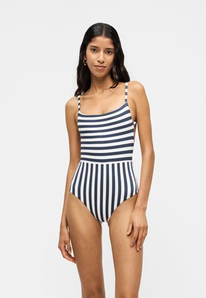 Femme aux longs cheveux foncés portant un maillot de bain une pièce rayé bleu marine et blanc avec des rayures horizontales sur le haut et verticales sur le bas.