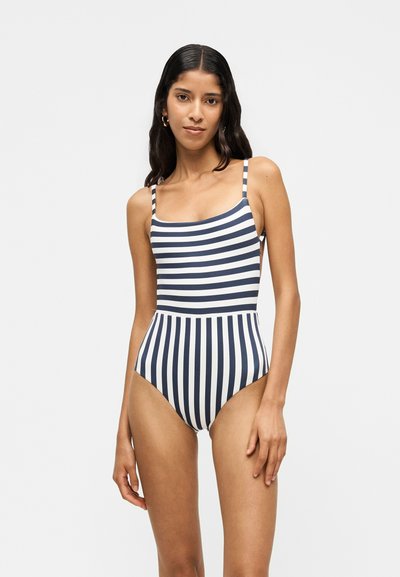 Femme aux longs cheveux foncés portant un maillot de bain une pièce rayé bleu marine et blanc avec des rayures horizontales sur le haut et verticales sur le bas.