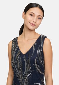 Robe navy avec un décolleté en V ornée de motifs de feuilles en argent et en paillettes. Tissu lisse avec un léger éclat, sans manches et silhouette ajustée.