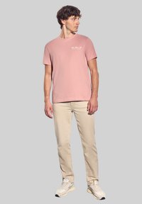 Světle růžové bavlněné tričko s krátkým rukávem, na kterém je bíly text. V kombinaci s béžovými rovnými kalhotami a světle zbarvenými teniskami.