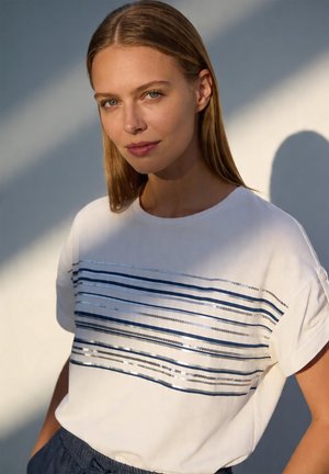 Junge Frau mit glatten blonden Haaren, die ein weißes T-Shirt mit horizontalen blauen und silbernen Streifen trägt, steht vor einem hellgrauen Hintergrund.