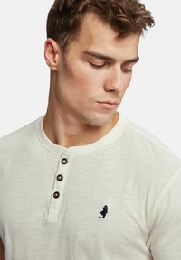 Kremkrāsas henley krekls, kas izgatavots no mīkstas auduma, ar pogām pa fronti un mazu izsistojumu figūriņu kreisajā krūtī.