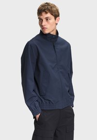 Jonge man die een marineblauwe jas met rits en hoge hals draagt, zwarte cargobroek, handen in de zakken, kijkt opzij.