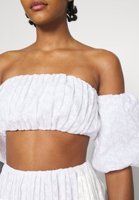 Top corto blanco con escote off-the-shoulder, textura fruncida y mangas abullonadas. Presenta un sutil patrón floral y una banda elástica en la cintura.